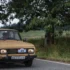 Old beige vintage car on a country road with a large tree in the background; three people visible through the windshield.