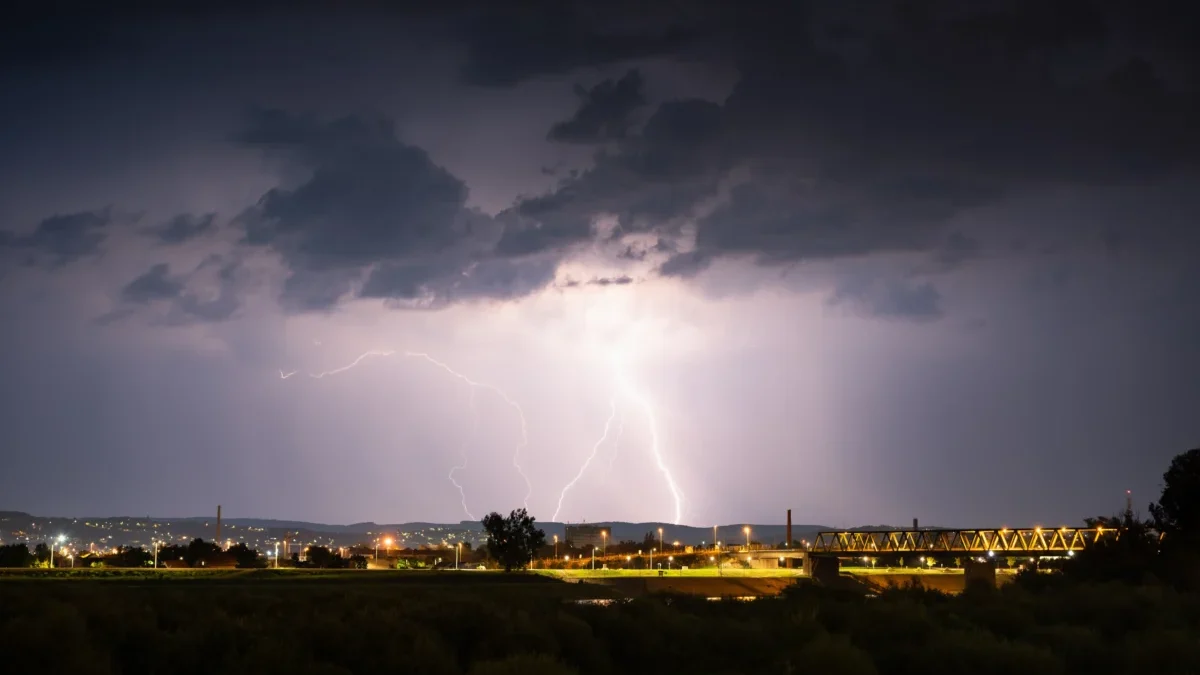 Nighttime storm over a city: bright lightning forks illuminate dark clouds above a lit bridge and distant buildings.