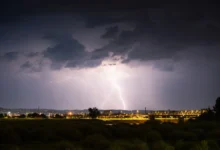 Nighttime storm over a city: bright lightning forks illuminate dark clouds above a lit bridge and distant buildings.