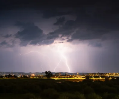 Nighttime storm over a city: bright lightning forks illuminate dark clouds above a lit bridge and distant buildings.