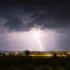 Nighttime storm over a city: bright lightning forks illuminate dark clouds above a lit bridge and distant buildings.