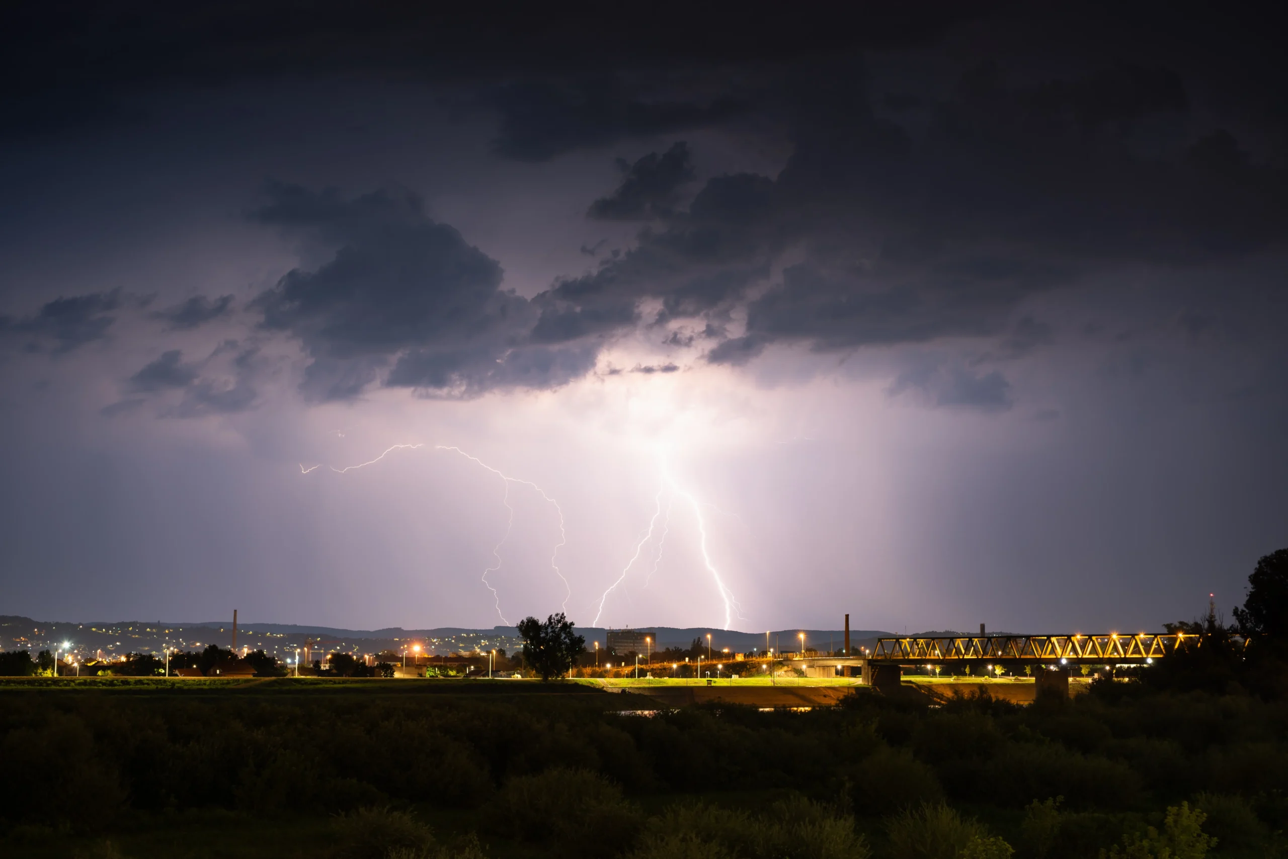 Nighttime storm over a city: bright lightning forks illuminate dark clouds above a lit bridge and distant buildings.