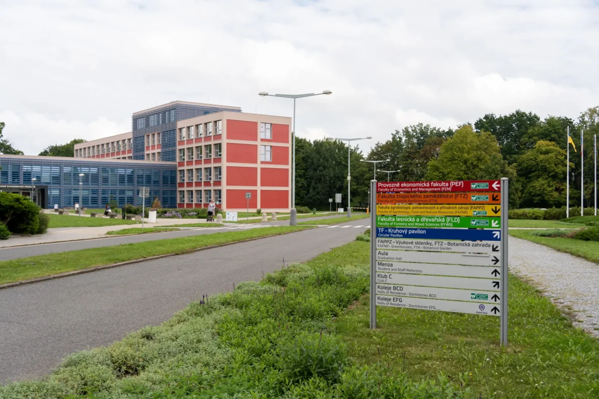 arealCZU | TVGURU.cz Barevný ukazatel s pokyny ke kampusu před moderními červenomodrými univerzitními budovami a stromy pod zataženou oblohou.