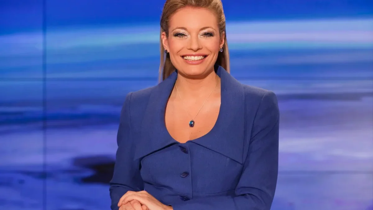 Professional female news anchor seated at a desk, wearing a blue blazer, smiling at the camera in a blue-toned studio backdrop with horizontal light bands.