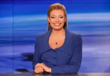 Professional female news anchor seated at a desk, wearing a blue blazer, smiling at the camera in a blue-toned studio backdrop with horizontal light bands.