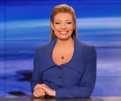 Professional female news anchor seated at a desk, wearing a blue blazer, smiling at the camera in a blue-toned studio backdrop with horizontal light bands.