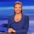 Professional female news anchor seated at a desk, wearing a blue blazer, smiling at the camera in a blue-toned studio backdrop with horizontal light bands.