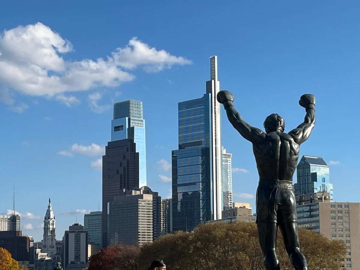 shutterstock_2702656073 1 | TVGURU.cz Bronze statue in foreground with arms raised, overlooking a modern city skyline under a blue sky.