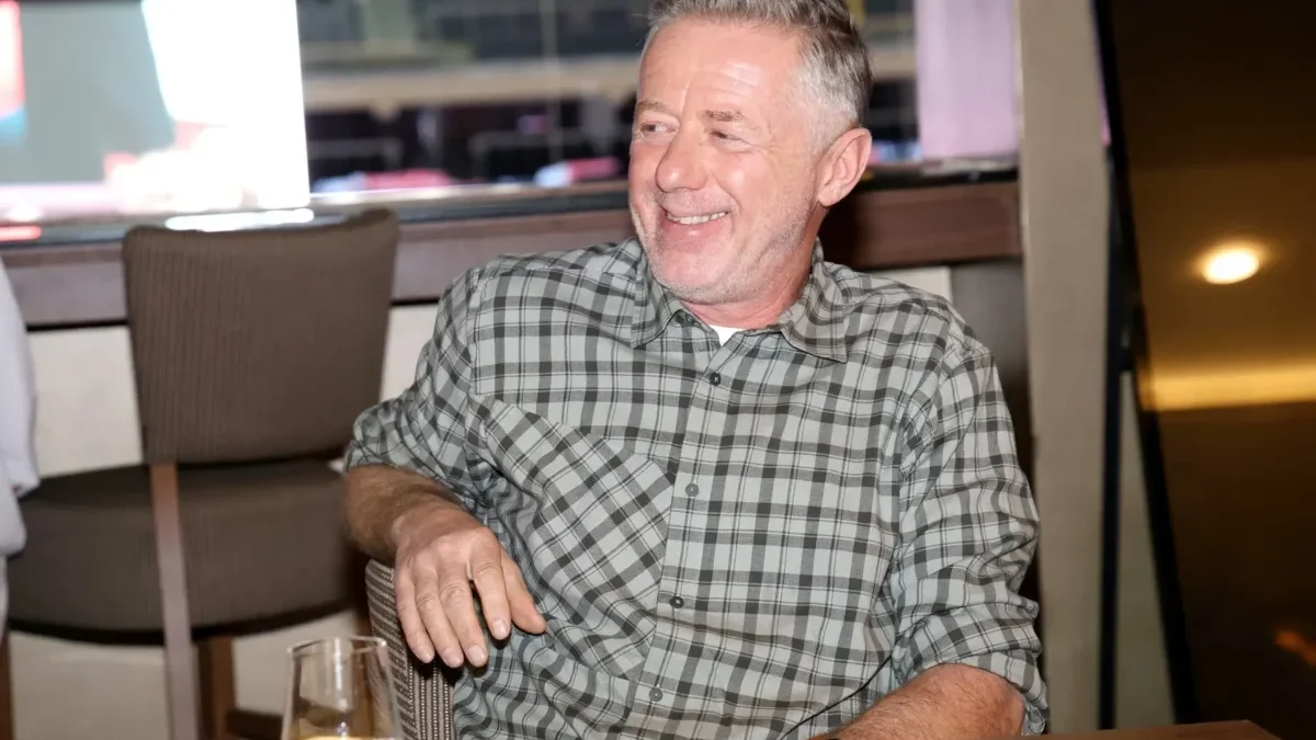 Smiling, middle-aged man with gray hair wearing a gray plaid shirt sits at a restaurant table, a glass of water with lemon in the foreground.