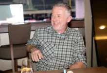 Smiling, middle-aged man with gray hair wearing a gray plaid shirt sits at a restaurant table, a glass of water with lemon in the foreground.