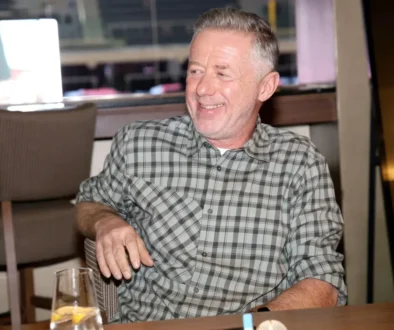 Smiling, middle-aged man with gray hair wearing a gray plaid shirt sits at a restaurant table, a glass of water with lemon in the foreground.