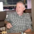 Smiling, middle-aged man with gray hair wearing a gray plaid shirt sits at a restaurant table, a glass of water with lemon in the foreground.
