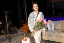 Smiling woman in a denim jacket and white outfit holds a bouquet of pink roses on a stage set, with a cake nearby.