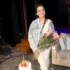 Smiling woman in a denim jacket and white outfit holds a bouquet of pink roses on a stage set, with a cake nearby.