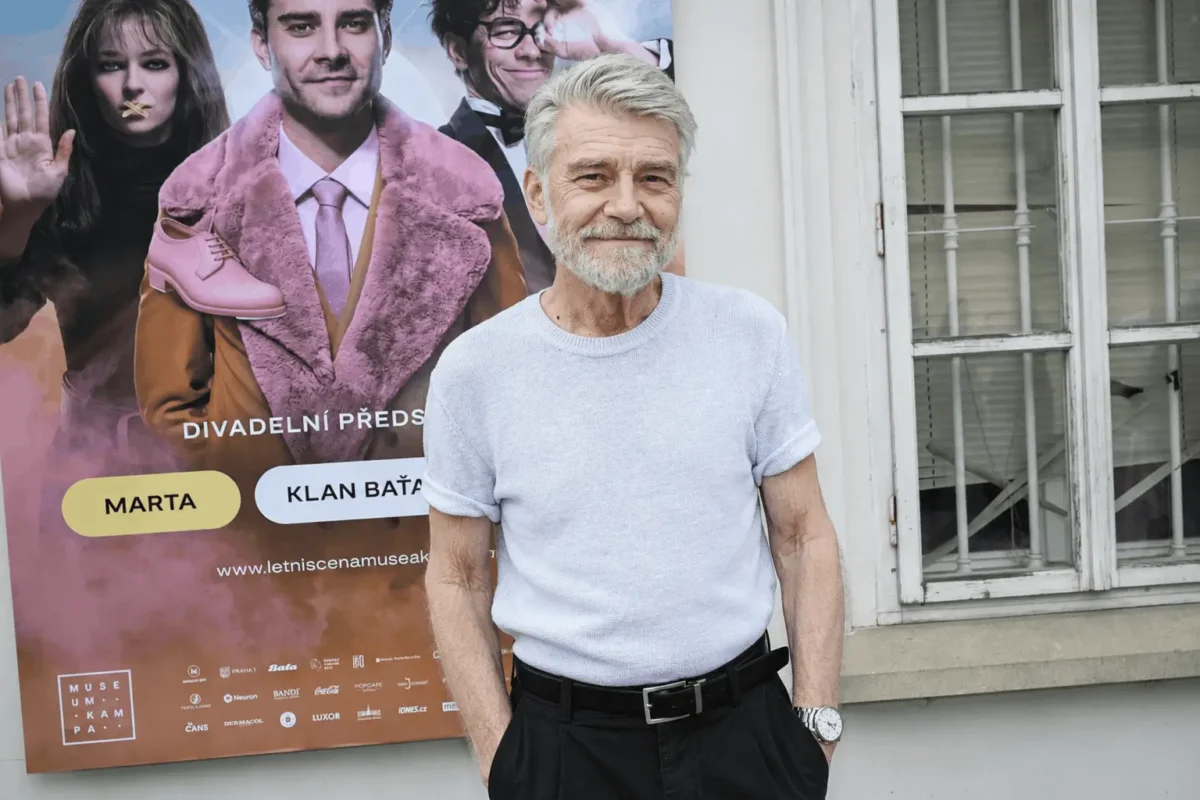 ChatGPT Image 1 7 2026 11_00_14 1 | TVGURU.cz Older man with gray hair and beard in a light blue knit shirt poses in front of a colorful poster featuring a pink shoe and stylish characters, beside a white window frame.