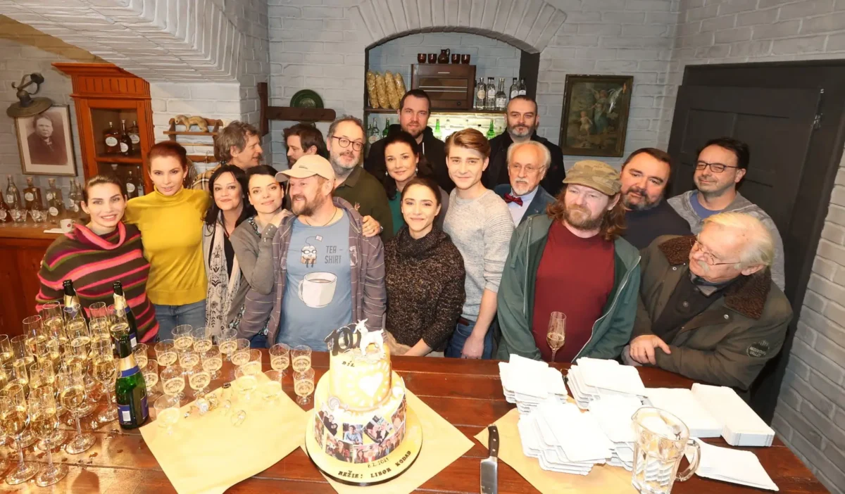profimedia-0589643968 1 | TVGURU.cz A group of people gathered in a cozy bar around a table with a two-tier birthday cake and champagne glasses, posing for a photo.