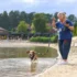 Woman in a blue top stands on a sandy lakeshore, holding a leash while a dog runs into the water beside her.