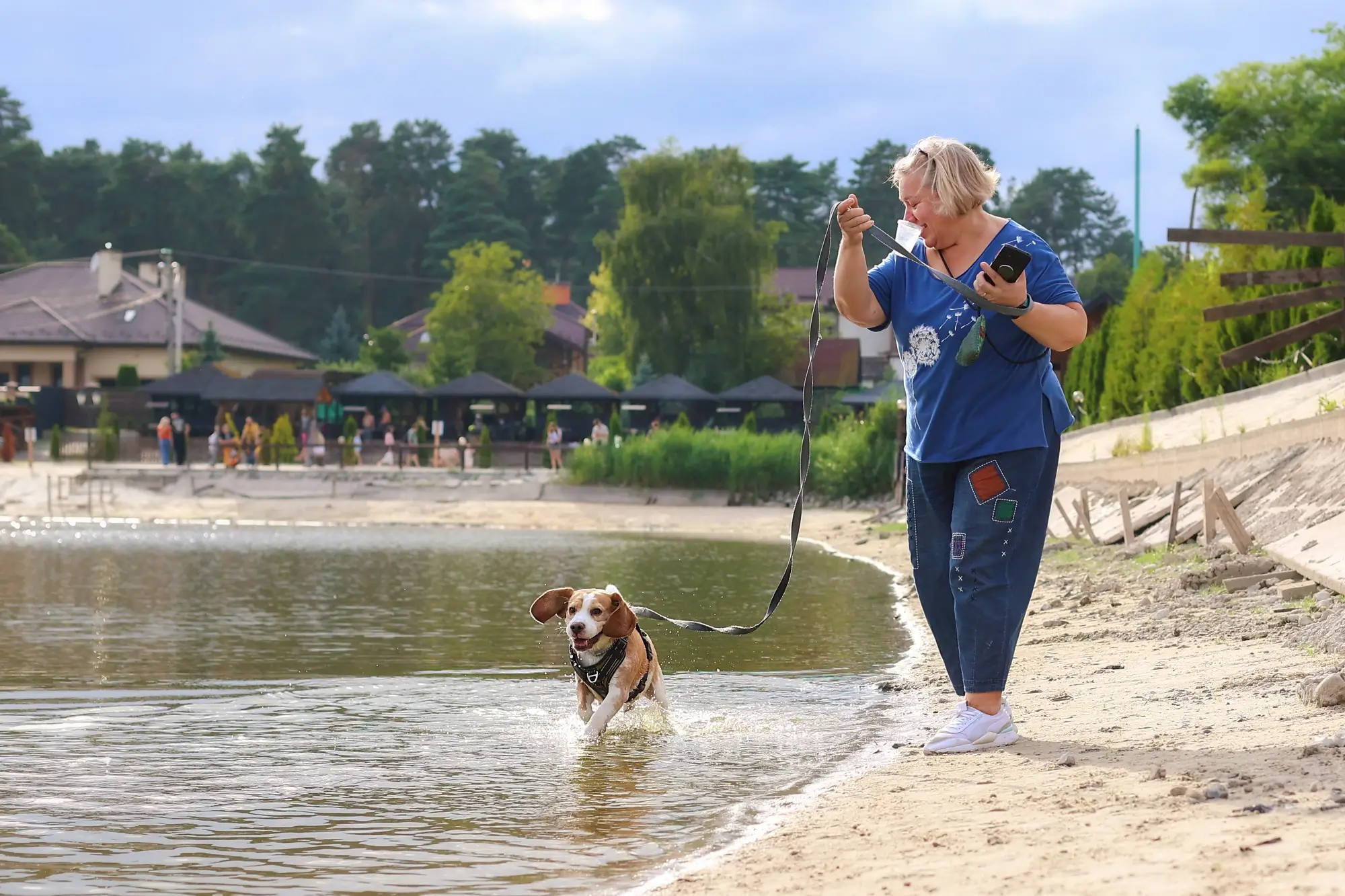 Woman in a blue top stands on a sandy lakeshore, holding a leash while a dog runs into the water beside her.