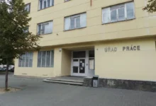 Yellow government building facade with 'ÚŘAD PRÁCE' sign above the entrance and glass doors; steps lead to the entry, tree on left side.