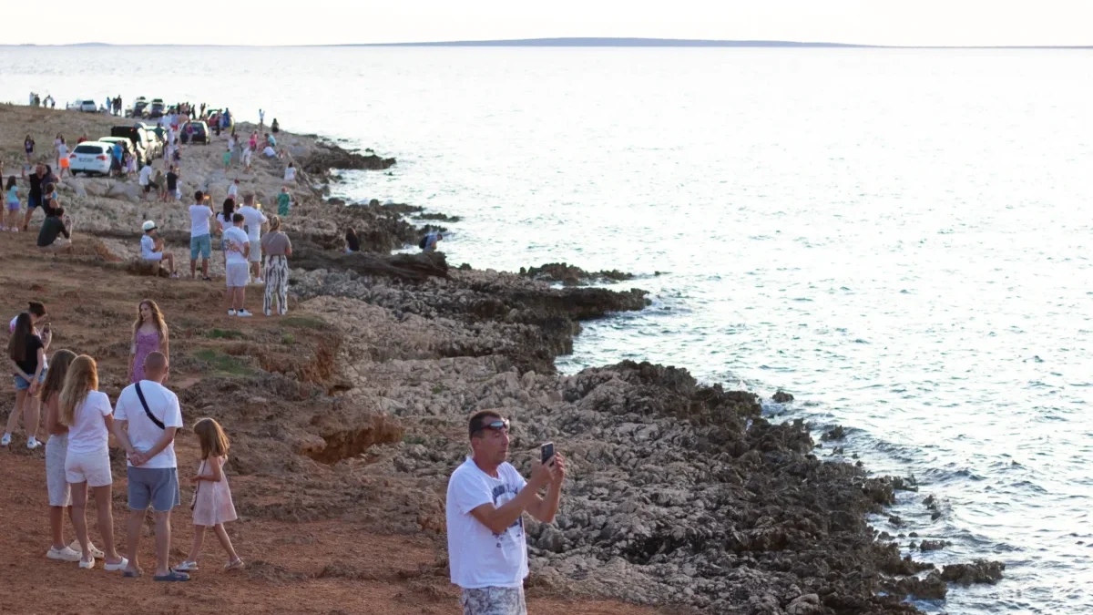 Crowded rocky shoreline with groups of people, cars parked along the coast, and the calm sea extending to the horizon.
