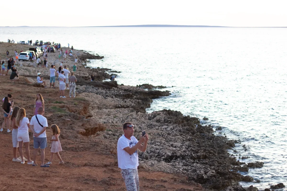 Crowded rocky shoreline with groups of people, cars parked along the coast, and the calm sea extending to the horizon.