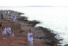 Crowded rocky shoreline with groups of people, cars parked along the coast, and the calm sea extending to the horizon.