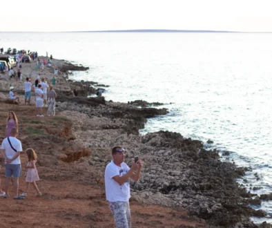 Crowded rocky shoreline with groups of people, cars parked along the coast, and the calm sea extending to the horizon.