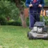 Person operating a gas-powered lawn mower on a green lawn with trees and a house in the background, in a backyard setting.
