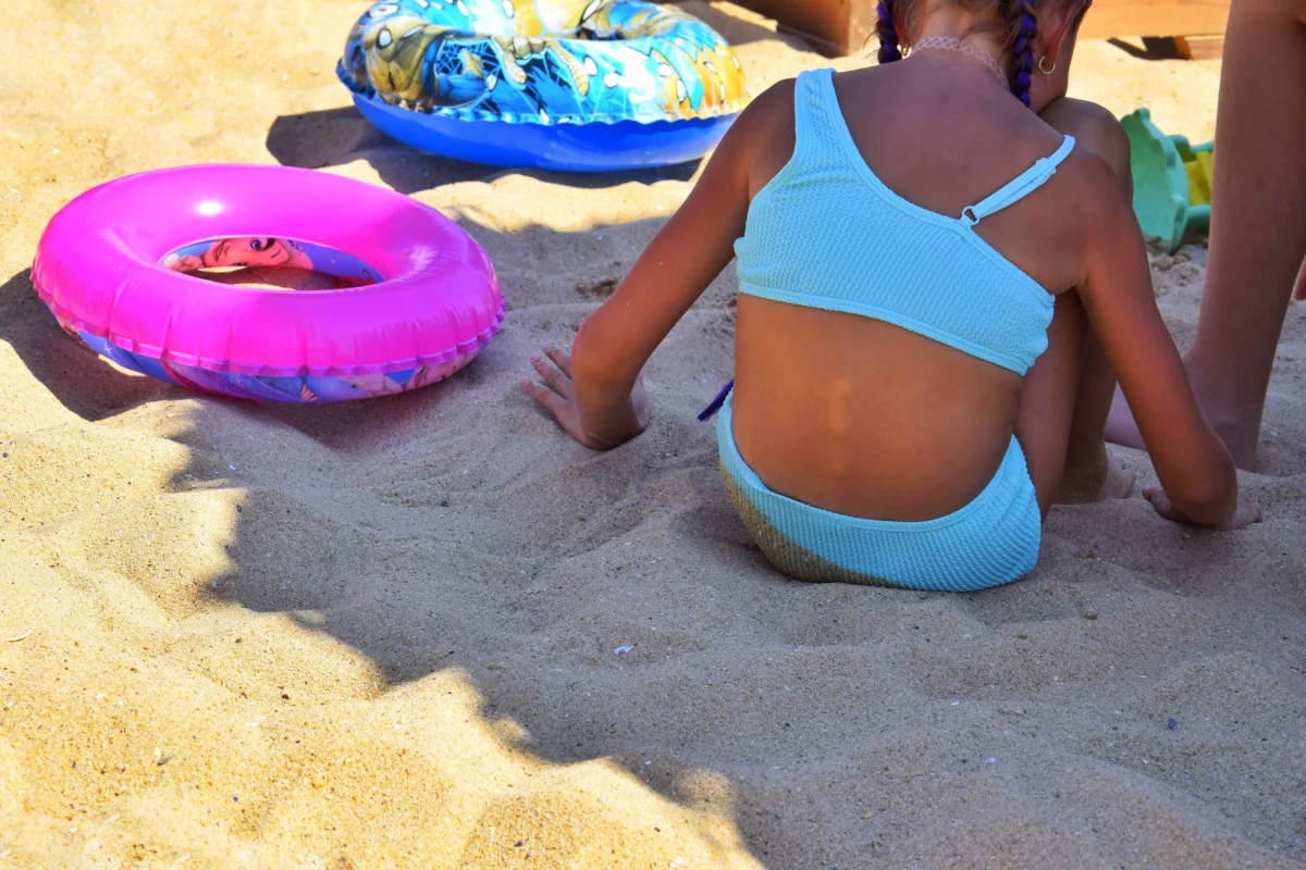 Children at the beach with bright inflatable swim toys, seen from behind a girl in a light blue swimsuit sitting on the sand.