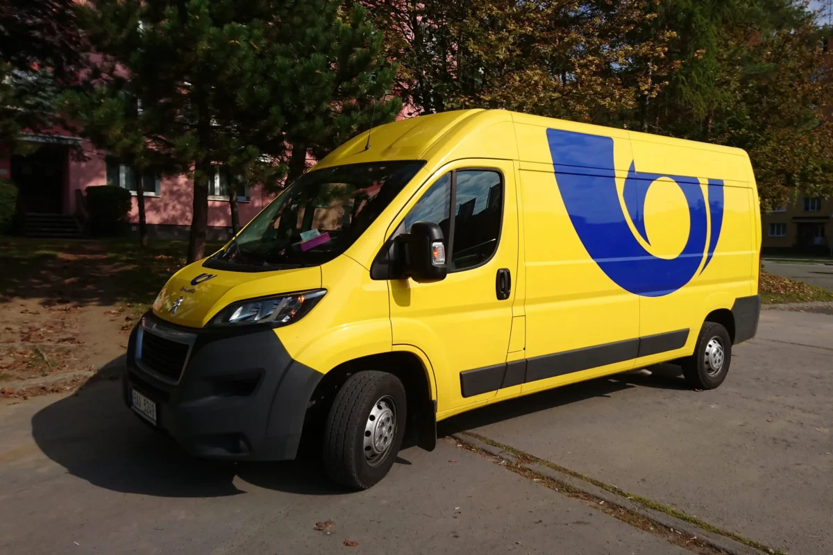 shutterstock_491429293 1 | TVGURU.cz Front-left view of a bright yellow delivery van with a blue emblem on its side, parked by trees and a pink building in the background.