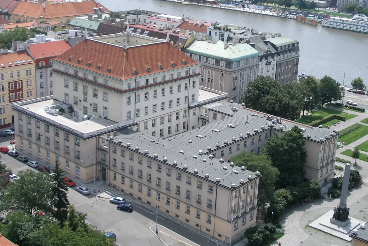 02 1 | TVGURU.cz Aerial view of a European city riverfront with red-tiled roofs, beige buildings, trees, and a statue in a foreground plaza.