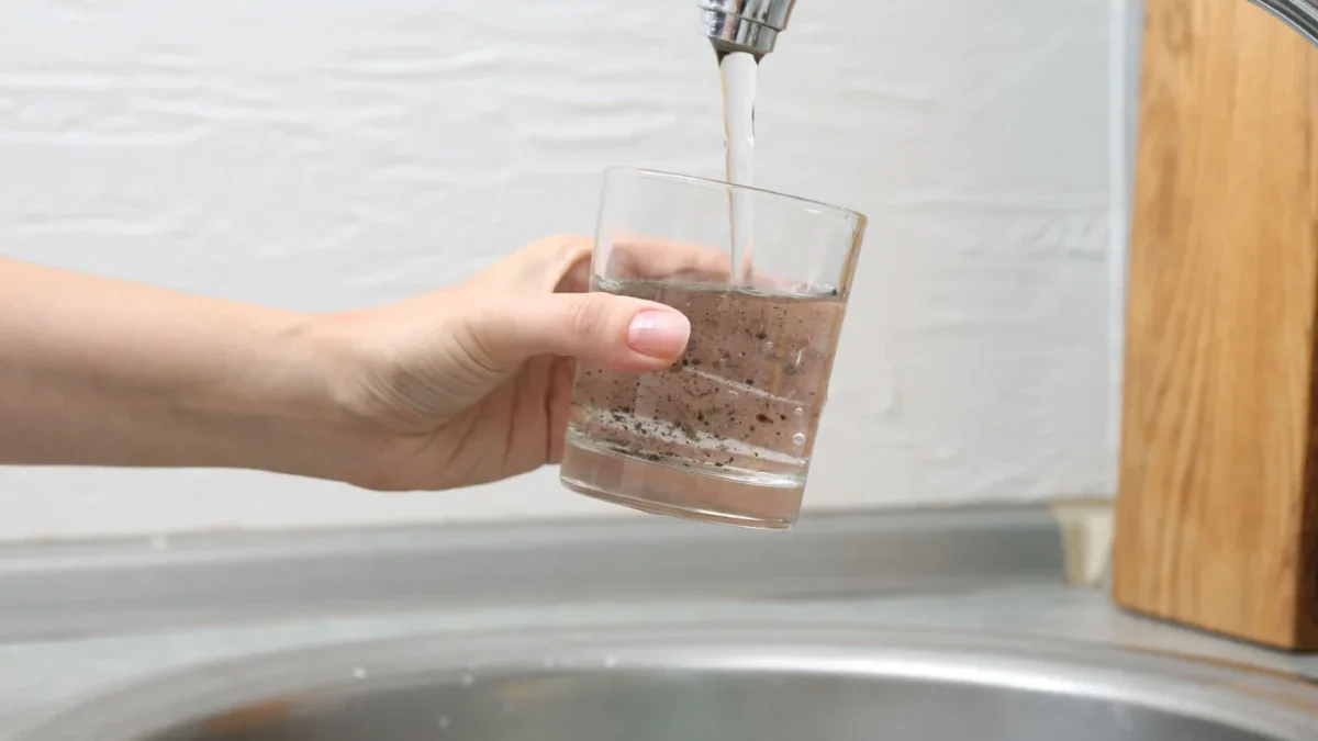 Person holds a glass under a running faucet, filling it with murky brown water.