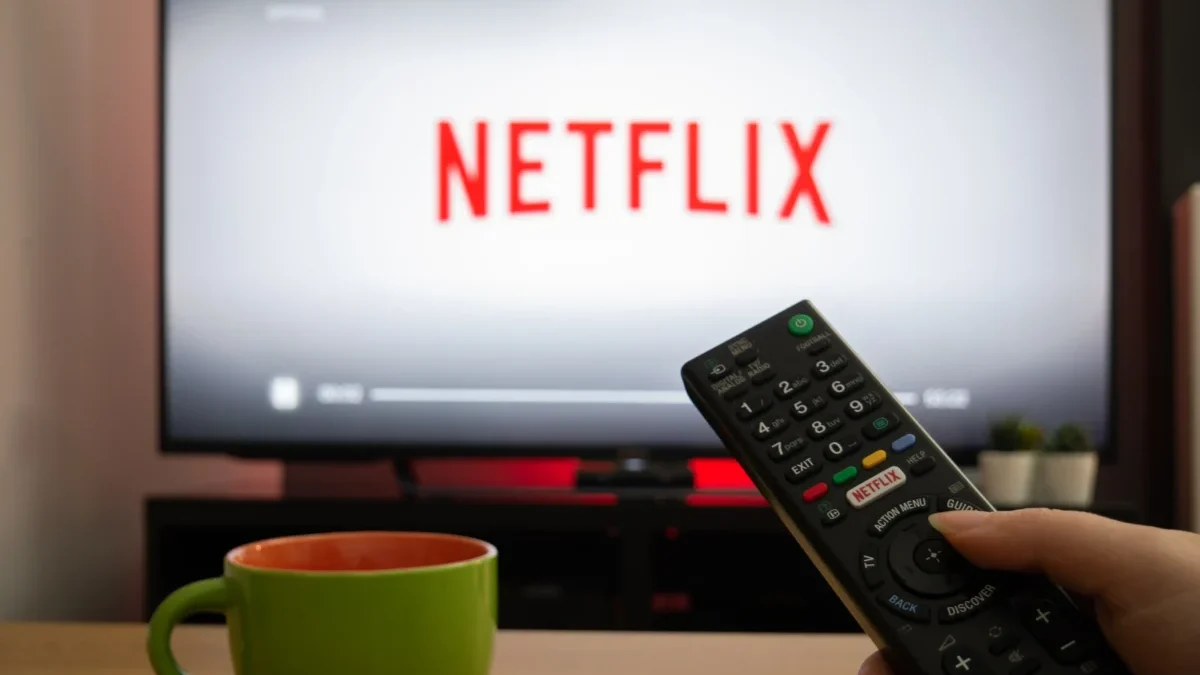 Close-up of a black TV remote in a hand, pointing toward a screen that displays the Netflix logo
