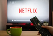 Close-up of a black TV remote in a hand, pointing toward a screen that displays the Netflix logo