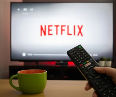 Close-up of a black TV remote in a hand, pointing toward a screen that displays the Netflix logo