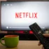 Close-up of a black TV remote in a hand, pointing toward a screen that displays the Netflix logo