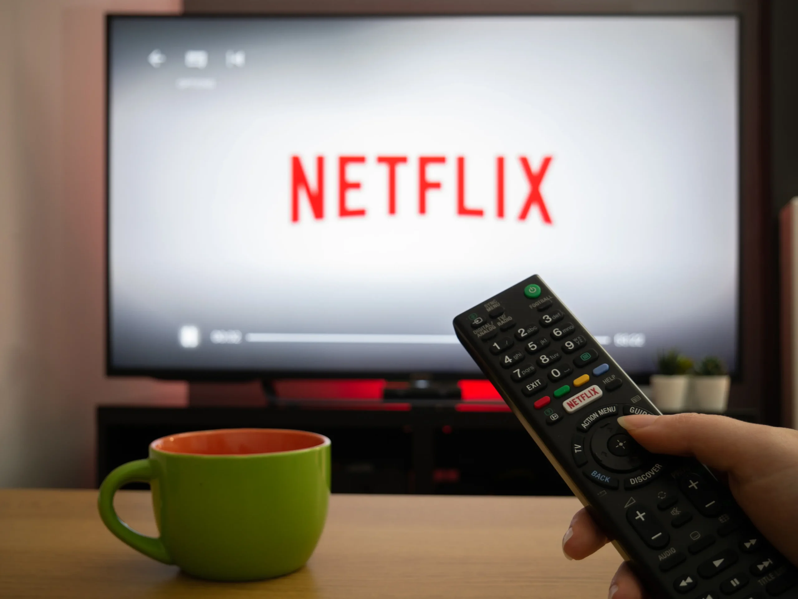 Close-up of a black TV remote in a hand, pointing toward a screen that displays the Netflix logo