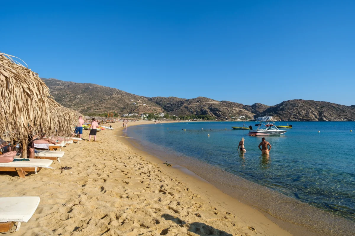 shutterstock_2737462281 1 | TVGURU.cz Sunny beach with straw umbrellas and loungers along the sandy shore, calm turquoise water and rocky hills in the distance.