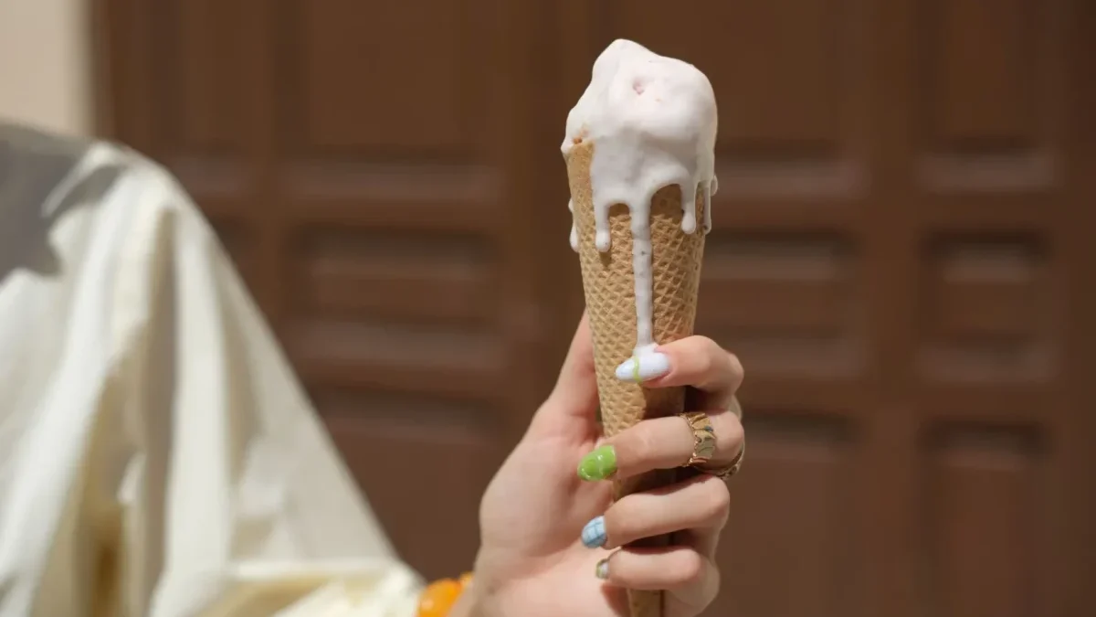 Person holding a vanilla ice cream cone with melted white ice cream dripping down the cone, bokeh background.