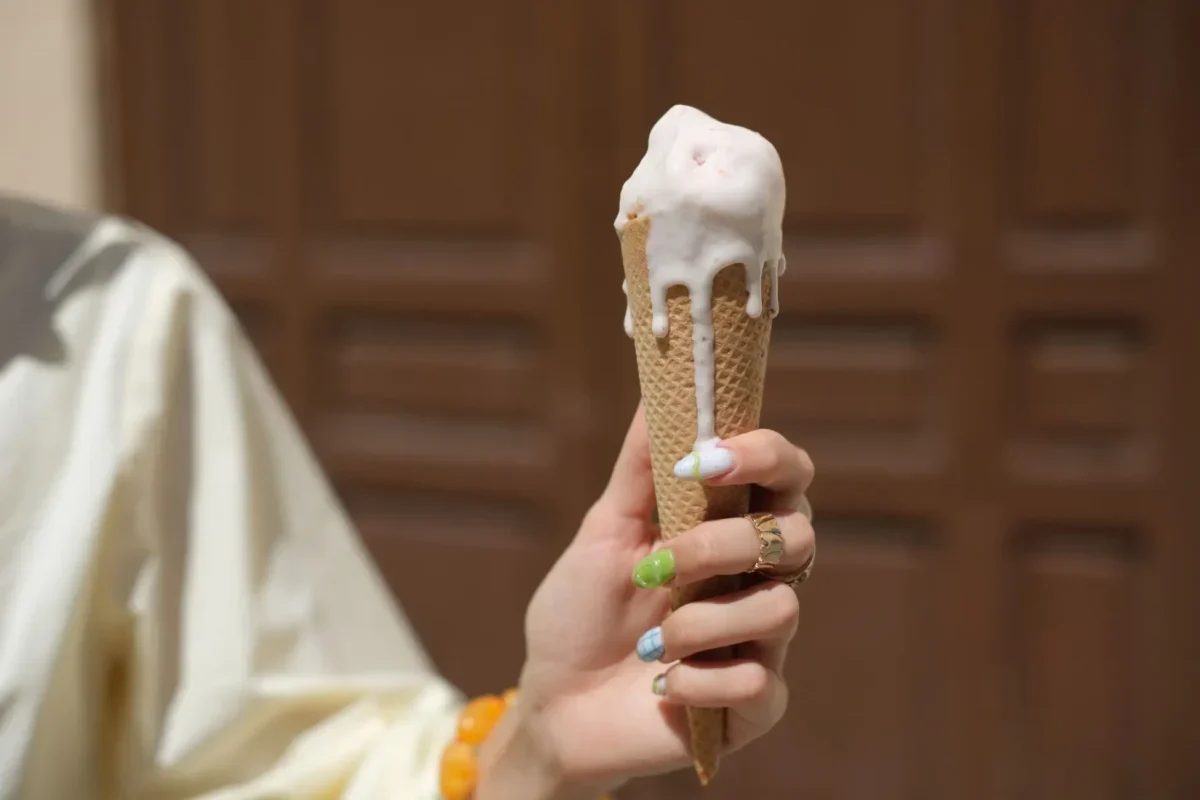 Person holding a vanilla ice cream cone with melted white ice cream dripping down the cone, bokeh background.