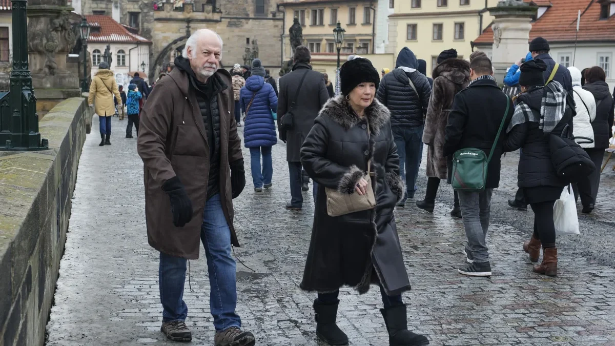 Crowded cobblestone street in a historic town with people wearing winter coats and fur, foreground man and woman looking toward the camera.