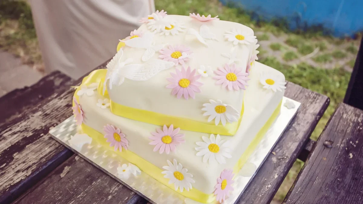 Two-tier white cake adorned with pink and white daisies on a wooden table outdoors. (Informative)