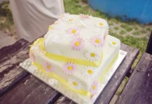 Two-tier white cake adorned with pink and white daisies on a wooden table outdoors. (Informative)