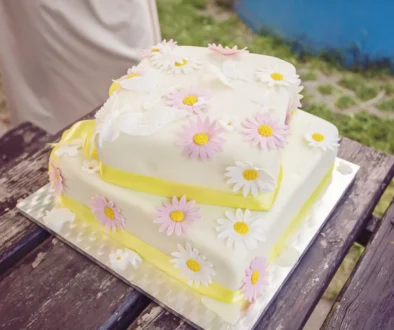 Two-tier white cake adorned with pink and white daisies on a wooden table outdoors. (Informative)