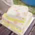 Two-tier white cake adorned with pink and white daisies on a wooden table outdoors. (Informative)