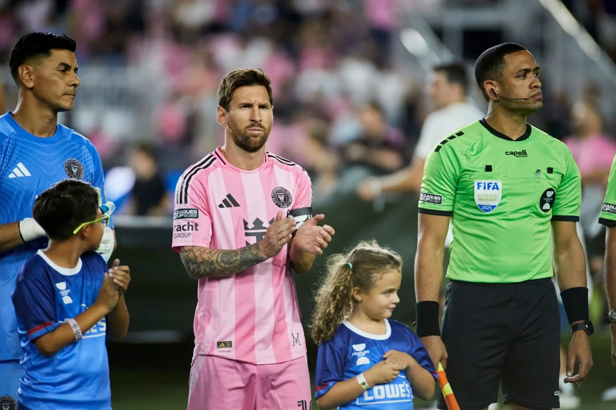 messi | TVGURU.cz Fotbalisté a rozhodčí stojí na hřišti před zápasem; hráč uprostřed má na sobě dres s růžovými pruhy, hráč vlevo je v modrém a rozhodčí vpravo v zeleném.
