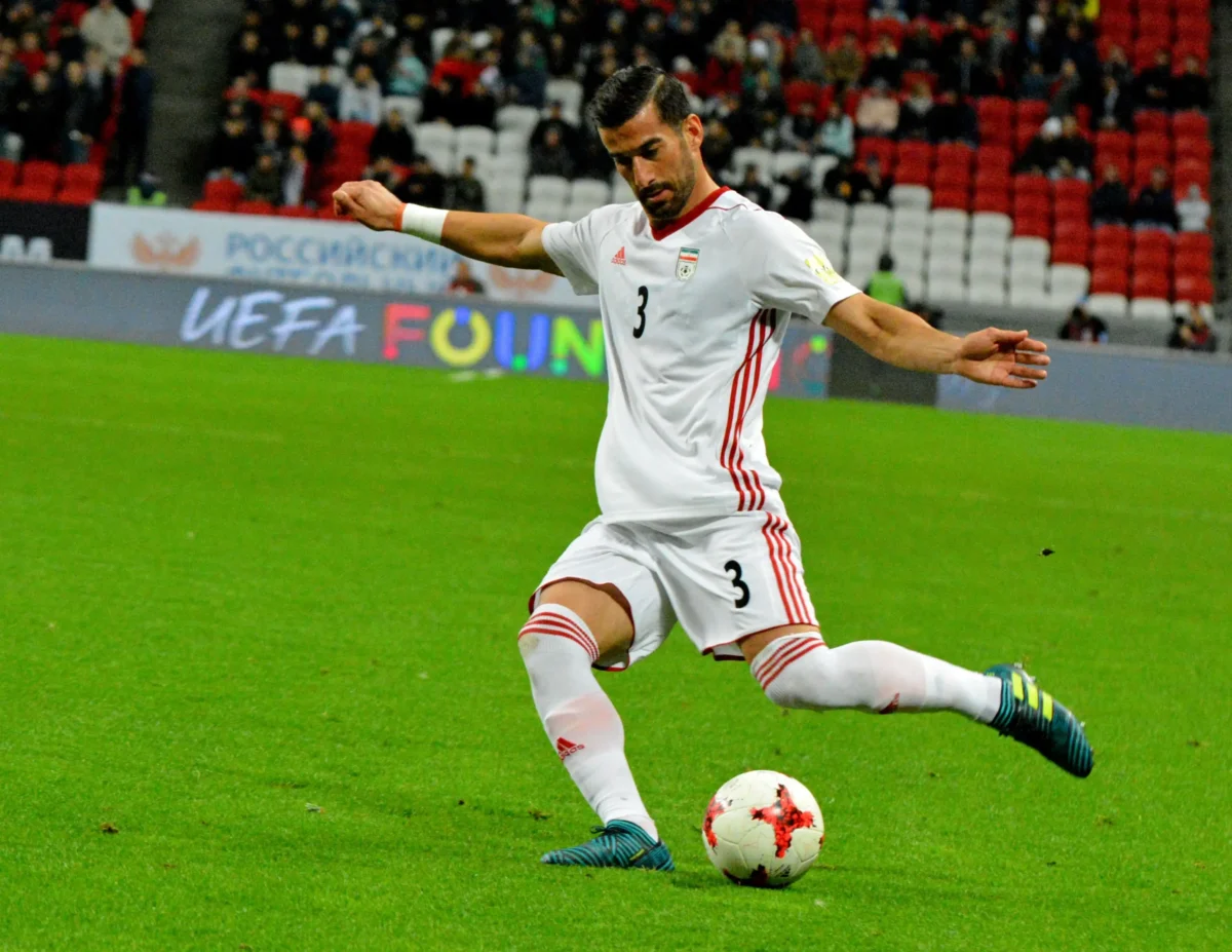 obrana | TVGURU.cz Fotbalista v bílém dresu Íránu s číslem 3 vede míč po zeleném hřišti, v pozadí jsou tribuny a diváci.