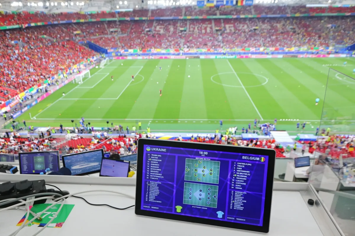 sledovani | TVGURU.cz Pohled z komentátorského stanoviště na zaplněný stadion s fotbalovým hřištěm; obrazovky tabletů zobrazují podrobnosti o sestavách pro zápas Ukrajina – Belgie.