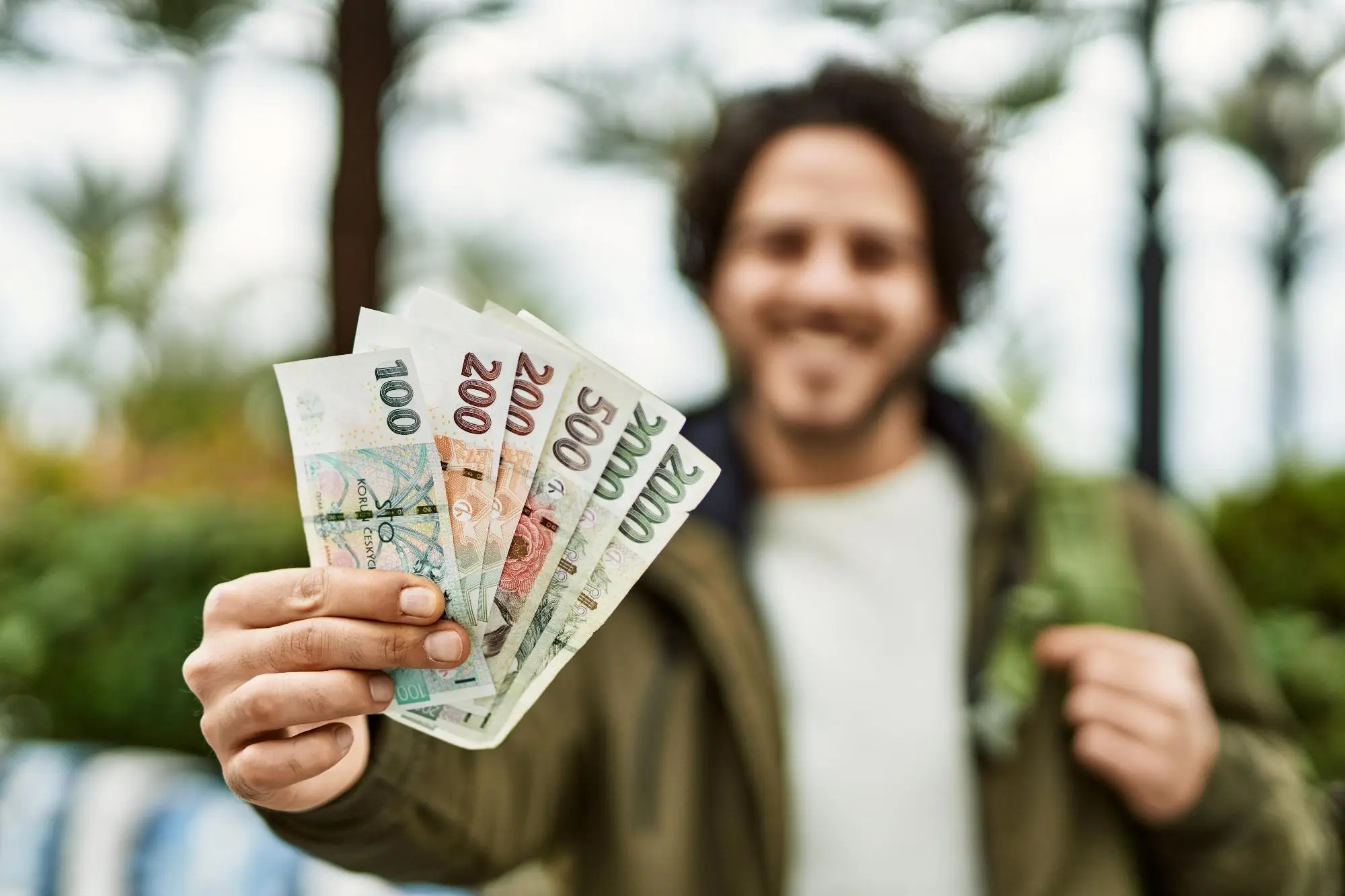 Superdávka vše usnadní | TVGURU.cz Person holds a fanned-out stack of euro banknotes toward the camera, smiling in an outdoor setting (background blurred).