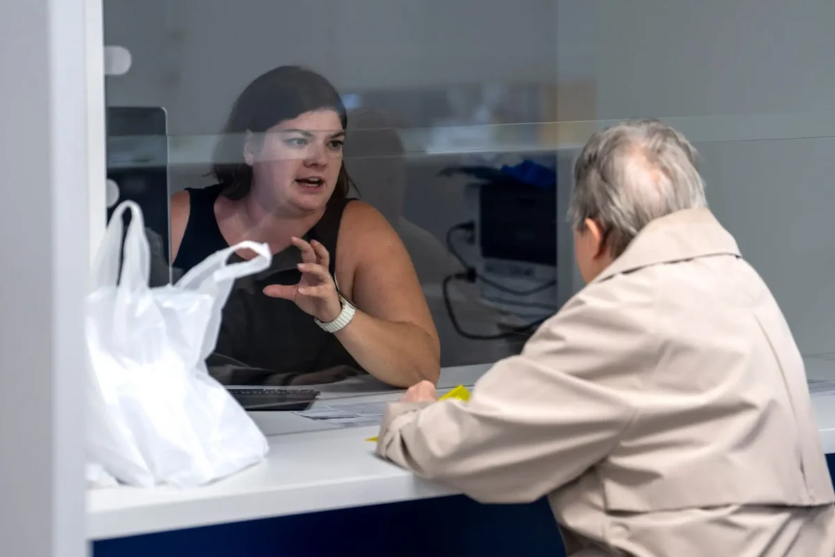 Woman behind a glass barrier speaks to an older man across a counter, gesturing with her hand as they converse.